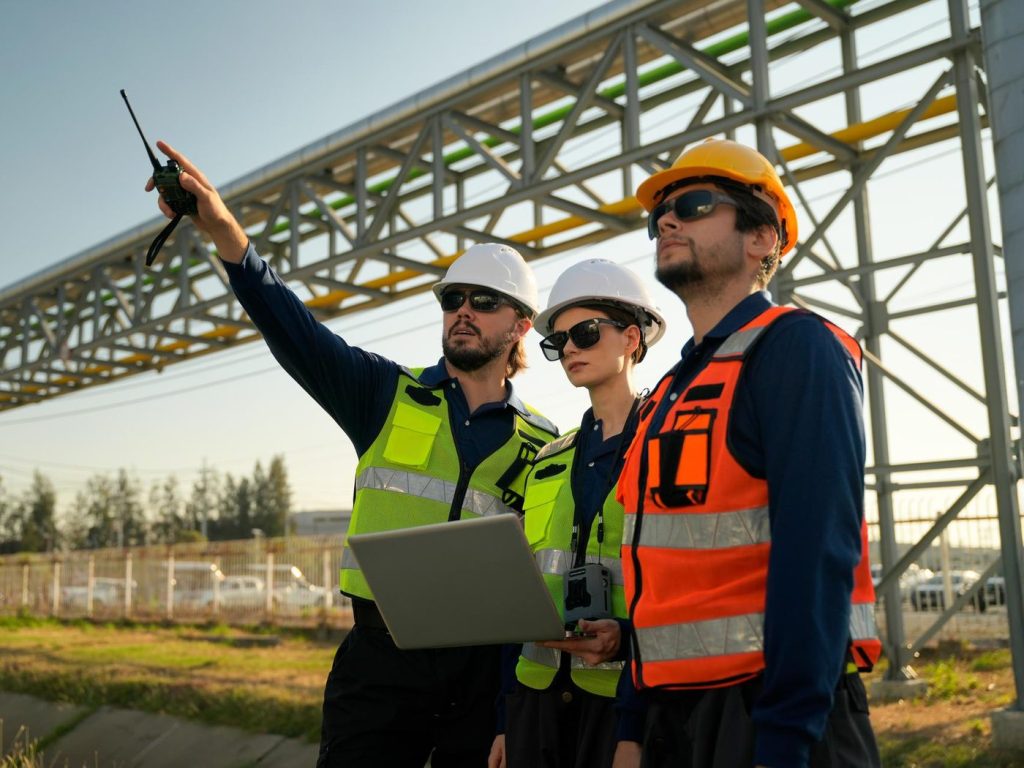 Tres especialistas con casco, lentes y chaleco reflectivo inspeccionan una instalación industrial con laptop y radio